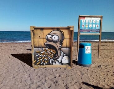 Homer Simpson vomitando colillas en la playa de Dénia llama mas la atención que el cartel de advertencia al lado.