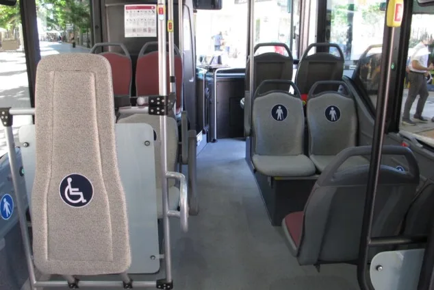 Imagen: Vista interior del autobús urbano de Bus Dénia que muestra el espacio adaptado para sillas de ruedas con respaldo acolchado y los asientos grises reservados con pictogramas para personas con discapacidad o movilidad reducida