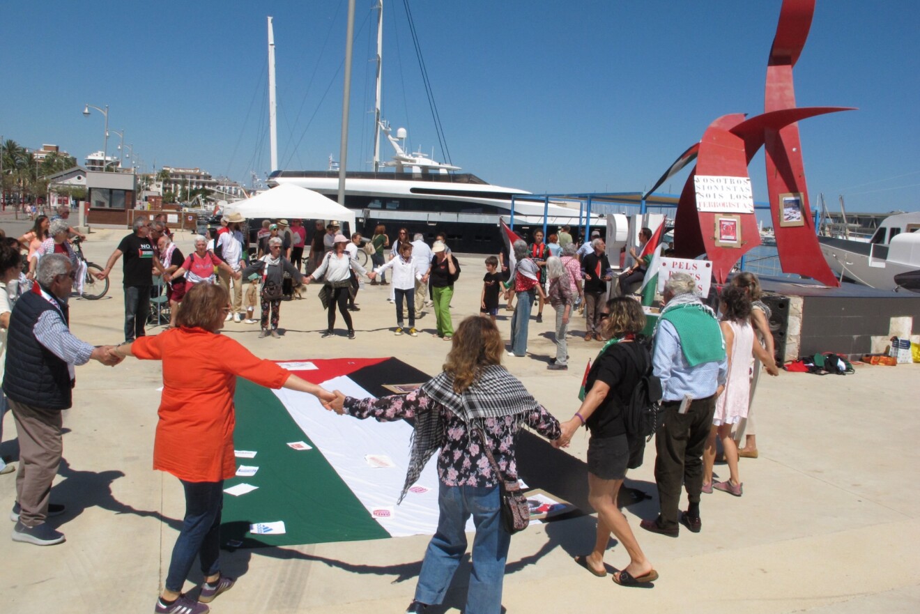 Un círculo de ciudadanos rodea la bandera palestina durante la jornada reivindicativa en el puerto de Dénia
