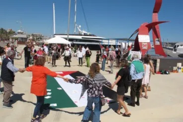 Un círculo de ciudadanos rodea la bandera palestina durante la jornada reivindicativa en el puerto de Dénia