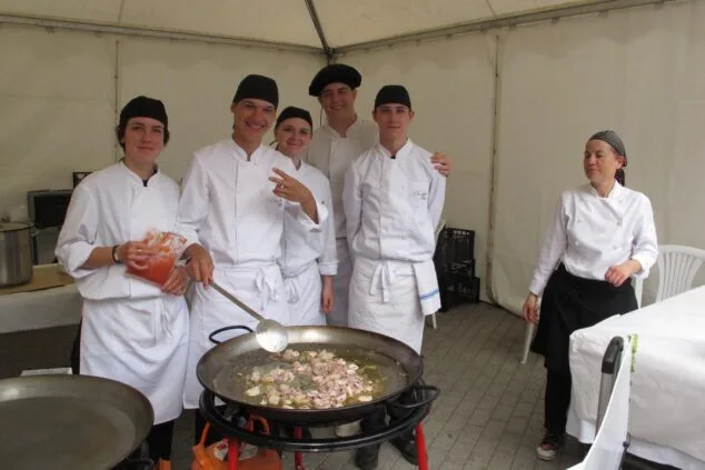 Dénia saca la Fira de l’Estudiantat 2026 a la calle por primera vez 5 Grupo de estudiantes de cocina con uniforme preparando comida en una paellera grande durante una feria al aire libre.