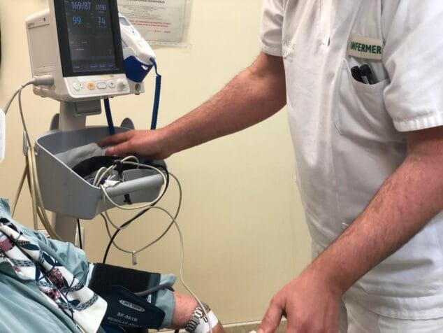 Eine Krankenschwester in weißer Uniform mit der Aufschrift „NURSE“ misst den Blutdruck eines Patienten, dessen Arm mit einer Manschette an einen Vitalzeichenmonitor angeschlossen ist.