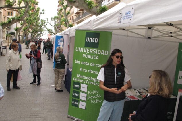 Stand de la UNED con una informadora y público en una feria educativa al aire libre en Dénia.