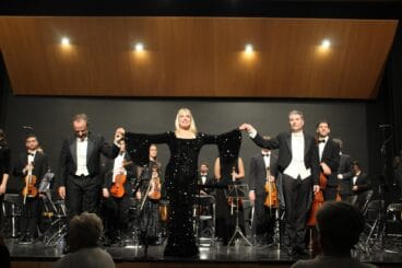 Irina Avramenko, Javier Palacios y Pere Molina saludan de la mano en el escenario tras la gala de ópera y zarzuela en Dénia con la orquesta al fondo.