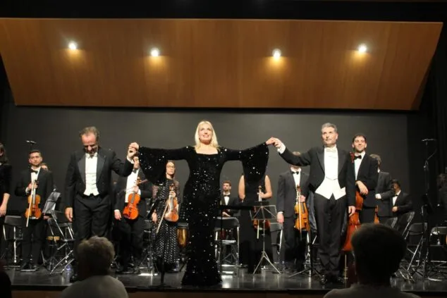 Irina Avramenko, Javier Palacios y Pere Molina saludan de la mano en el escenario tras la gala de ópera y zarzuela en Dénia con la orquesta al fondo.