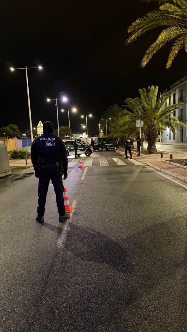 Operativo policial en Dénia: Un detenido y numerosas denuncias el sábado 2 Imagen: Un agente de la Policía Local de Dénia de espaldas en un control policial nocturno con patrullas y otros agentes al fondo, como parte de un operativo conjunto.