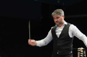 Fotografía del director de orquesta Pere Molina, vestido con chaleco negro, dirigiendo con la batuta en movimiento en un escenario oscuro.