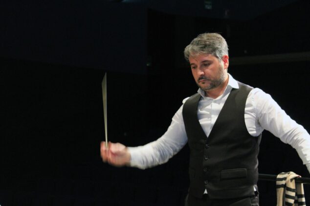 Imagen: Fotografía del director de orquesta Pere Molina, vestido con chaleco negro, dirigiendo con la batuta en movimiento en un escenario oscuro.