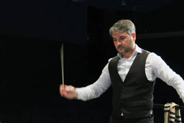 Fotografía del director de orquesta Pere Molina, vestido con chaleco negro, dirigiendo con la batuta en movimiento en un escenario oscuro.