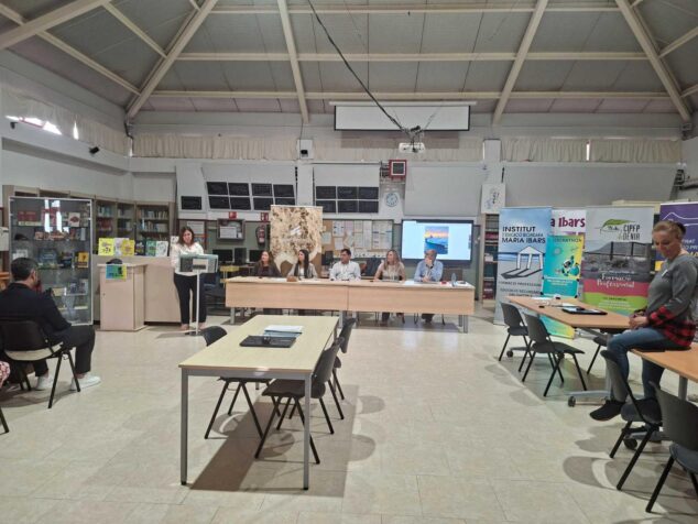Imagen: Vista general de la presentación del I Hackathon Intercentros Marina Alta en la biblioteca del IES Maria Ibars, con una oradora en un atril y una mesa con varios representantes.