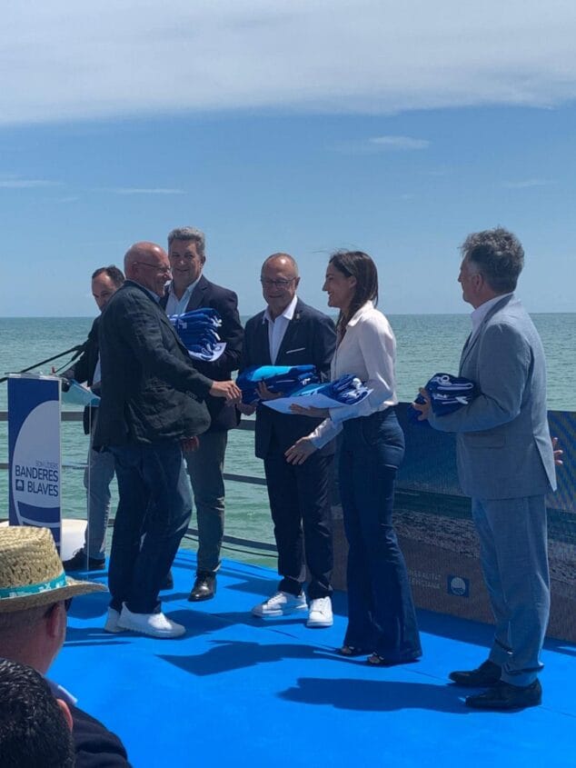 Imagen: Acto de entrega de las Banderas Azules con el alcalde y el concejal de Dénia en un escenario frente al mar.