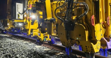 Primer plano de maquinaria ferroviaria amarilla realizando trabajos de bateo en las vías del TRAM d'Alacant durante la noche.