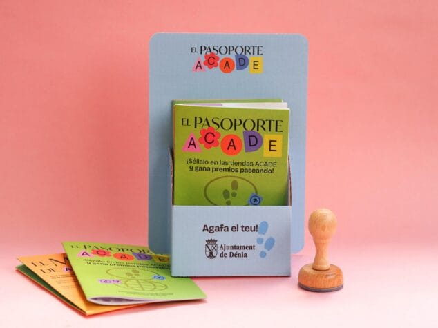 Expositor azul con folletos verdes de 'El Pasoporte ACADE', un sello de madera y más folletos sobre fondo rosa.