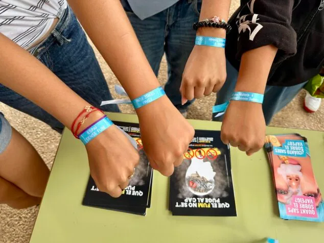 La verdad sobre vapers y cachimbas que Dénia está contando a sus estudiantes 1 Imagen: Primer plano de cuatro brazos de jóvenes con pulseras azules de la campaña "Espai Sense Fum" y folletos informativos sobre una mesa verde.