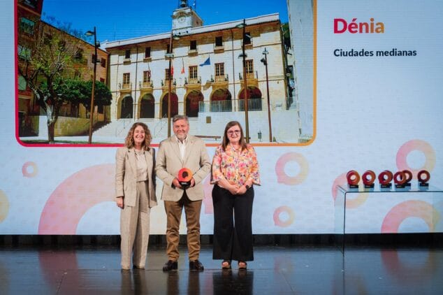 Imagen: El alcalde de Dénia, Rafa Carrió, y dos representantes del ayuntamiento sostienen el premio "Ciudad de la Ciencia y la Innovación" en un escenario con una pantalla de fondo que dice "Dénia".