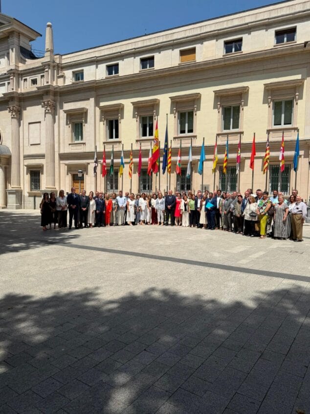 Imagen: Un grupo numeroso de personas posa frente a la fachada de un edificio institucional con banderas de España y comunidades autónomas bajo un sol brillante.