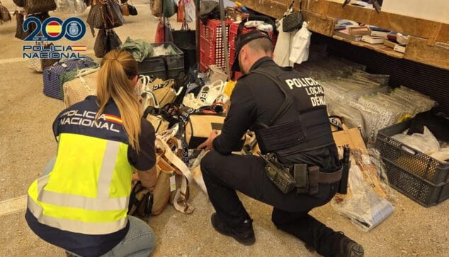 Cae una red de falsificaciones en Dénia: 12 detenidos y miles de artículos incautados 3 Imagen: Dos agentes de policía, uno de la Policía Nacional y otro de la Local de Denia, arrodillados inspeccionando bolsos y otros objetos en el suelo.