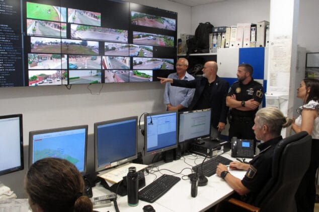 Imagen: Autoridades y policías en el centro de control de Dénia observando una gran pantalla con múltiples imágenes del sistema de cámaras de seguridad, mientras un hombre señala una de las vistas.
