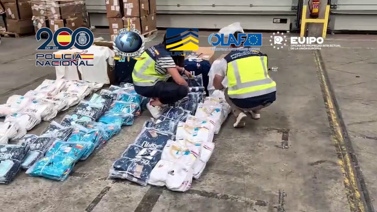 Procesamiento masivo de material incautado en un almacén, con gran cantidad de cajas apiladas y abiertas llenas de camisetas deportivas