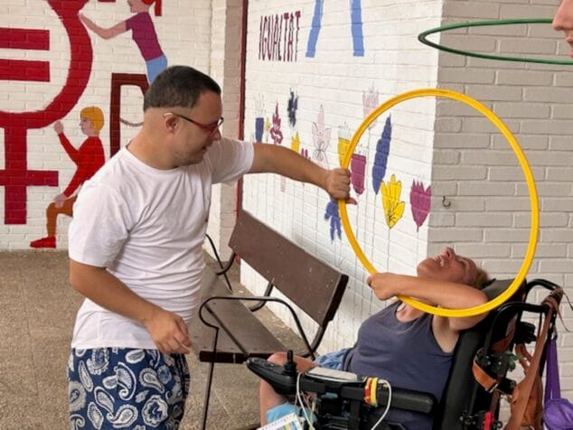 Condenados al Bordillo arranca su escuela de verano inclusiva con más de 50 participantes 1 Imagen: Un hombre con síndrome de Down sostiene un aro amarillo frente a una mujer en silla de ruedas durante una actividad.