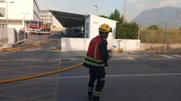 Declarado un incendio de vegetación en Dénia que moviliza a un amplio dispositivo de emergencias 8 Un bombero con casco y uniforme camina llevando una garrafa de agua, con un camión de bomberos y humo de un incendio forestal al fondo en Dénia.