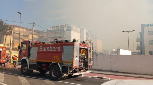 Declarado un incendio de vegetación en Dénia que moviliza a un amplio dispositivo de emergencias 9 Camión de bomberos rojo con la palabra Bombers aparcado en una calle de Dénia con humo en el cielo al fondo debido a un incendio de vegetación.