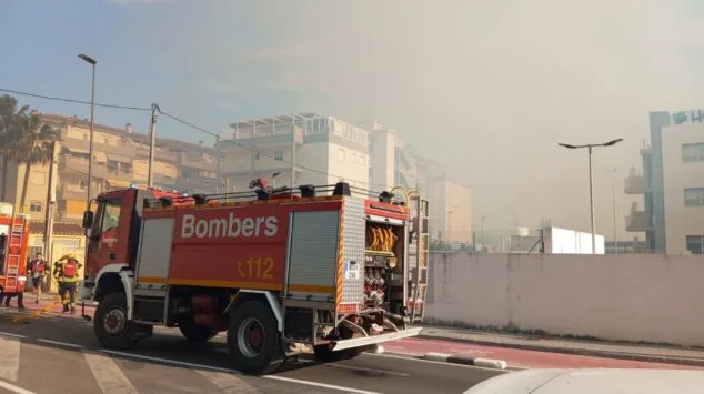 Declarado un incendio de vegetación en Dénia que moviliza a un amplio dispositivo de emergencias 9 Camión de bomberos rojo con la palabra Bombers aparcado en una calle de Dénia con humo en el cielo al fondo debido a un incendio de vegetación.