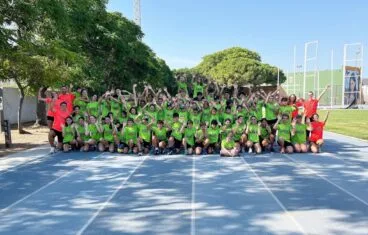 Numeroso grupo de niños y monitores con camisetas verdes posando en una pista de atletismo.