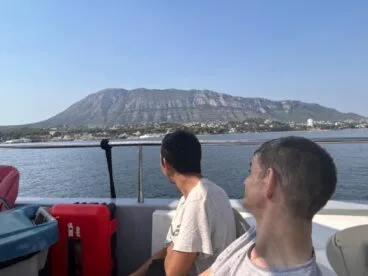 Condenados al Bordillo arranca su escuela de verano inclusiva con más de 50 participantes 59 Dos hombres en la cubierta de un barco mirando hacia la costa y el monte Montgó durante una actividad de verano.