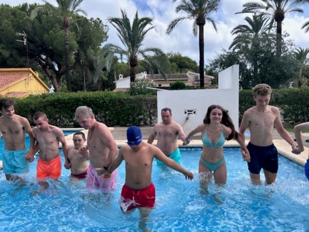 Condenados al Bordillo arranca su escuela de verano inclusiva con más de 50 participantes 5 Imagen: Grupo de participantes y monitores de Condenados al Bordillo cogidos de la mano en una piscina durante una actividad de verano.