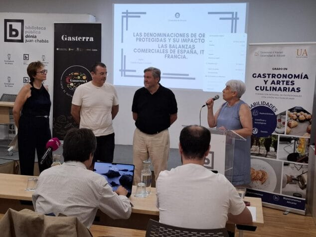 Dénia, epicentro de la innovación gastronómica con la defensa de los TFG de la UA 1 Imagen: Juana Jordá habla con micrófono durante la defensa de un TFG de Gastronomía, con una pantalla de presentación y otros asistentes.