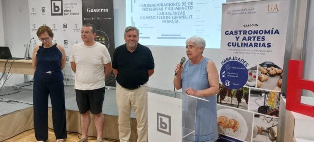 Dénia, epicentro de la innovación gastronómica con la defensa de los TFG de la UA 3 Imagen: Una mujer habla en un atril durante una presentación académica con una pantalla de proyección y otras tres personas de pie detrás.
