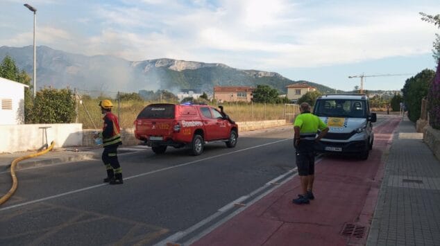 Declarado un incendio de vegetación en Dénia que moviliza a un amplio dispositivo de emergencias 10 Un bombero, personal de Protección Civil y vehículos de emergencia en una carretera de Dénia, con humo de un incendio visible al fondo.