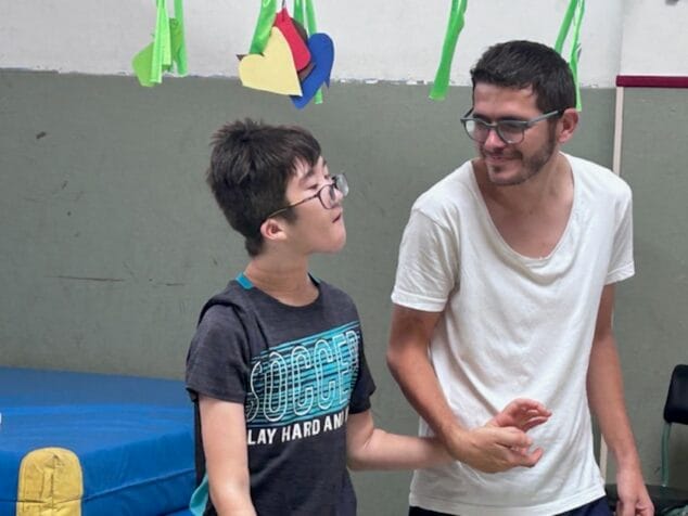 Condenados al Bordillo arranca su escuela de verano inclusiva con más de 50 participantes 2 Imagen: Un monitor sonríe mientras sostiene la muñeca de un participante joven con gafas en un gimnasio durante una actividad de la Escuela de Verano.