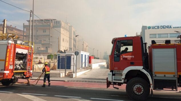 Declarado un incendio de vegetación en Dénia que moviliza a un amplio dispositivo de emergencias 2 Imagen: Dos camiones de bomberos de Alicante y un bombero trabajando en una calle de Dénia, con humo de incendio y el edificio del hospital HCB al fondo.