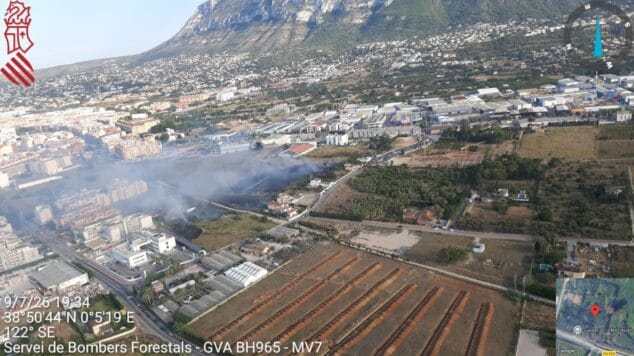 Declarado un incendio de vegetación en Dénia que moviliza a un amplio dispositivo de emergencias 1 Imagen: Vista aérea muestra una columna de humo saliendo de una zona de vegetación y campos cerca de una zona urbana en Dénia, con una montaña al fondo.