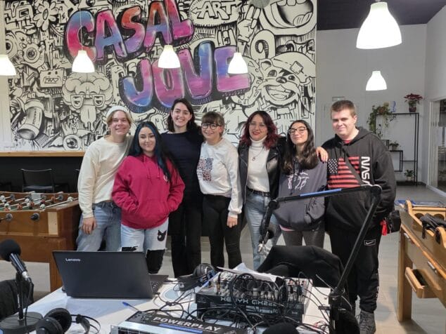 Jóvenes de Dénia debaten sobre salud mental, ocio y vivienda en el exitoso 'Pòdcast Jove' 2 Imagen: Fotografía de grupo de ocho jóvenes de pie detrás de una mesa con equipos de grabación de podcast como micrófonos y una mesa de mezclas, en una sala con un mural de grafiti que dice 'CASAL JOVE'.