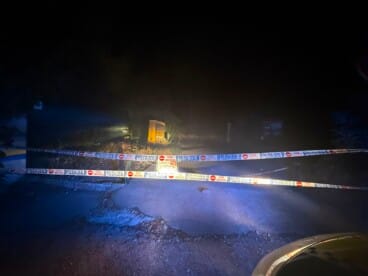 Cinta de la Policía Local corta el paso a un acceso durante la noche, iluminado por los focos de un vehículo, con señalización y vegetación al fondo.