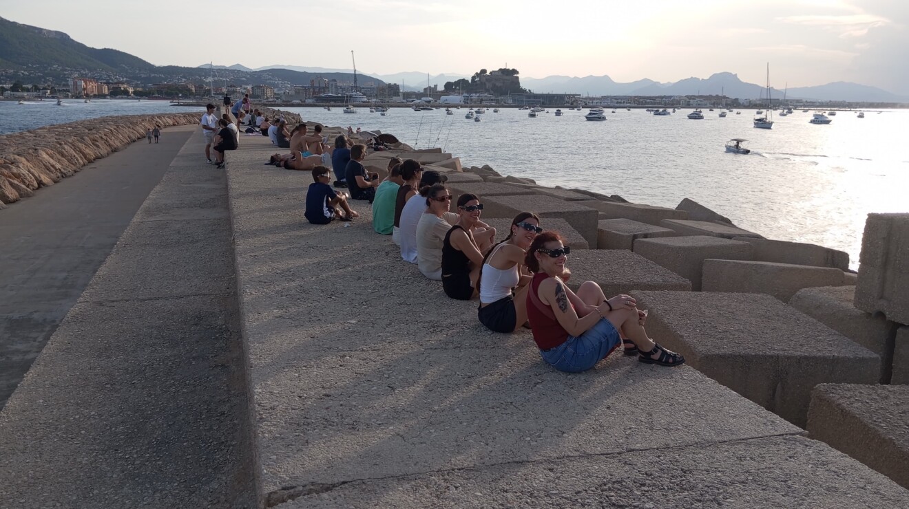 Personas esperando en las escolleras del puerto, con el Castell de Dénia al fondo y las gafas puestas