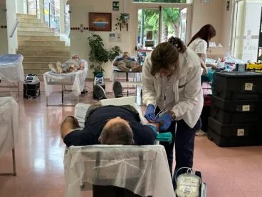 Una enfermera extrayendo sangre a un hombre en una camilla durante una jornada de donación, con otros donantes y personal al fondo.