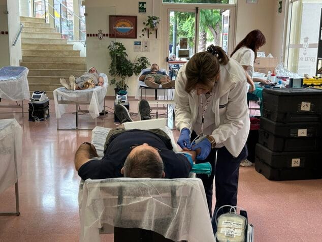 Una enfermera extrayendo sangre a un hombre en una camilla durante una jornada de donación, con otros donantes y personal al fondo.