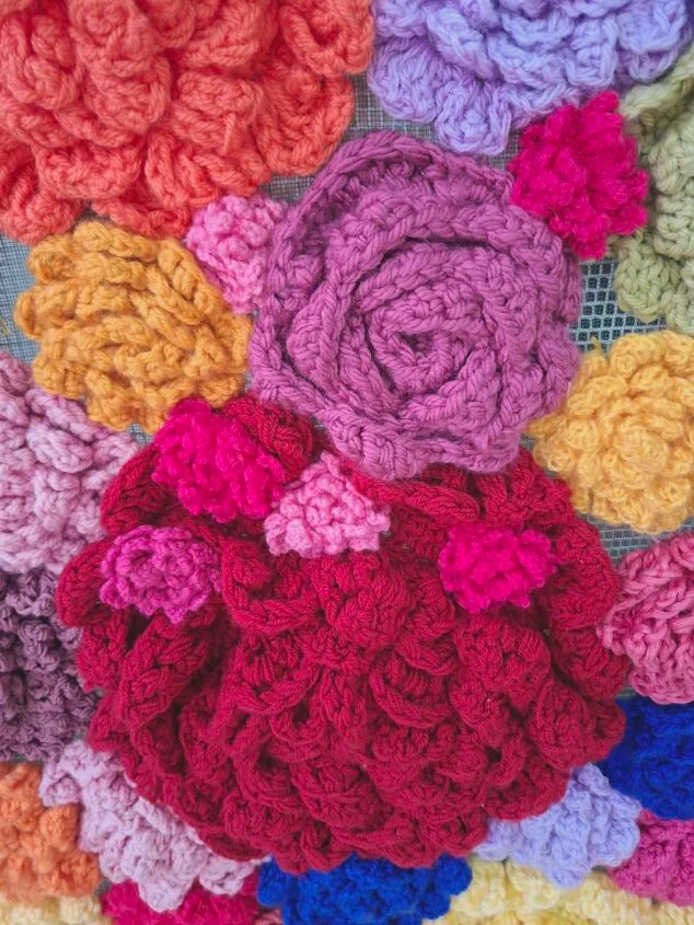 Imagen: Primer plano de una obra de arte textil compuesta por numerosas flores de ganchillo en colores vibrantes como rojo, rosa, naranja, morado y amarillo sobre una malla.
