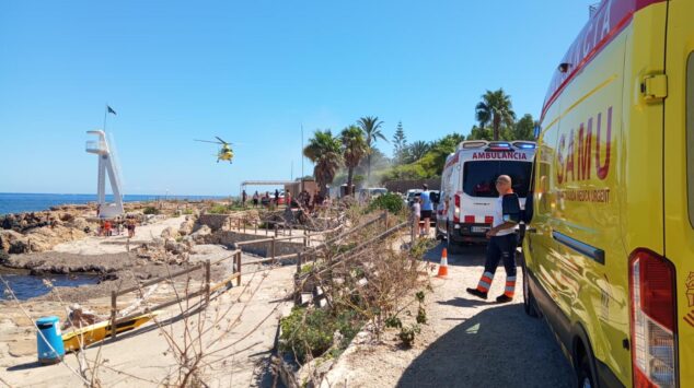 Fallece un hombre de 70 años tras sufrir un golpe en la cabeza en una playa de Dénia 5 Ambulancias SAMU y personal de emergencias en una zona costera rocosa con un helicóptero de rescate en el aire.
