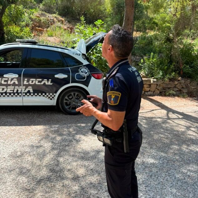 La Policía Local de Dénia localiza a dos senderistas perdidos en el Montgó con un dron 2 Imagen: Un agente de la Policía Local de Dénia maneja el controlador de un dron, mirando hacia arriba, junto a un coche patrulla en una zona rural.