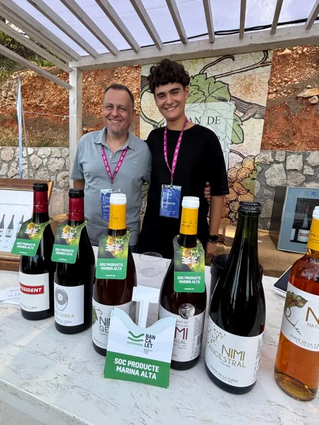 El secreto de la cocina local de la Marina Alta, desvelado en el D*na Festival 2 Imagen: Joan de la Casa y otro representante en su stand del D*na Festival con botellas de vino y el sello Bancalet.