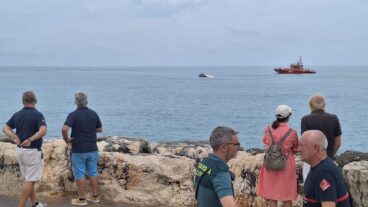 Varias personas, incluidos un guardia civil y un bombero, observan desde un espigón un velero volcado y una embarcación de rescate en el mar en Denia.