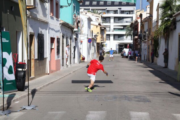 La «Setmana de la Pilota» llega a las calles de Dénia, a las aulas y dará voz a las mujeres del deporte 1 Imagen: Un jugador con camiseta roja golpea una pelota de vaqueta en una partida de pelota valenciana en una calle de Dénia con público al fondo.