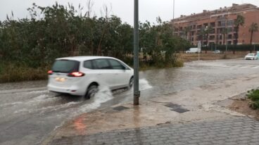 Dénia suspende las actividades al aire libre ante el aviso naranja por lluvias y tormentas 8 Un coche blanco circula por una calle inundada, levantando agua a su paso bajo un cielo gris.