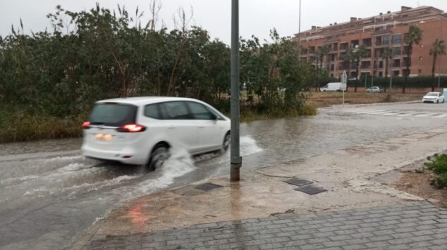 Un coche blanco circula por una calle inundada, levantando agua a su paso bajo un cielo gris.