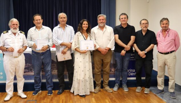 «Save the ocean» de Mapi Gómez, gana el primer premio de la 8ª edición de 'Esculturas frente al mar' de Dénia 3 Imagen: Siete personas posan de pie durante la entrega de premios. Tres de ellas, en el centro, sostienen diplomas. Un hombre con uniforme blanco está a la izquierda. Detrás hay una cortina azul.
